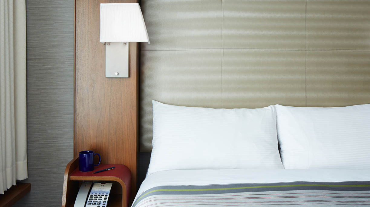 A close-up of a bed with white pillows and a striped blanket, with a nightstand and lamp beside it.