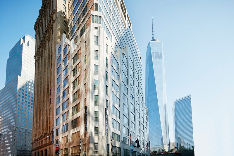 A large building with many windows stands next to other skyscrapers in a city, with a clear blue sky above.