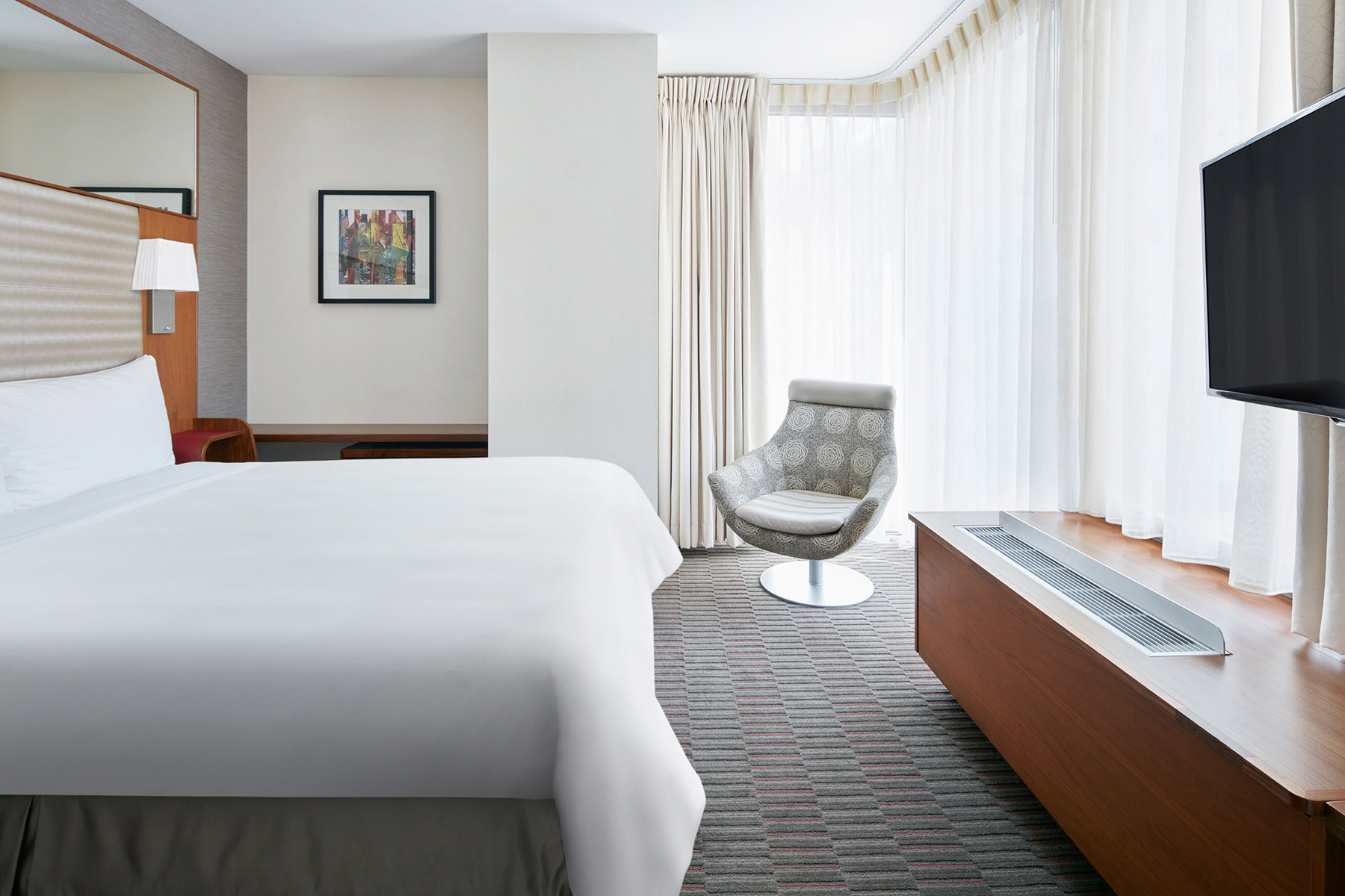 A hotel room with a bed, chair, TV, and window.