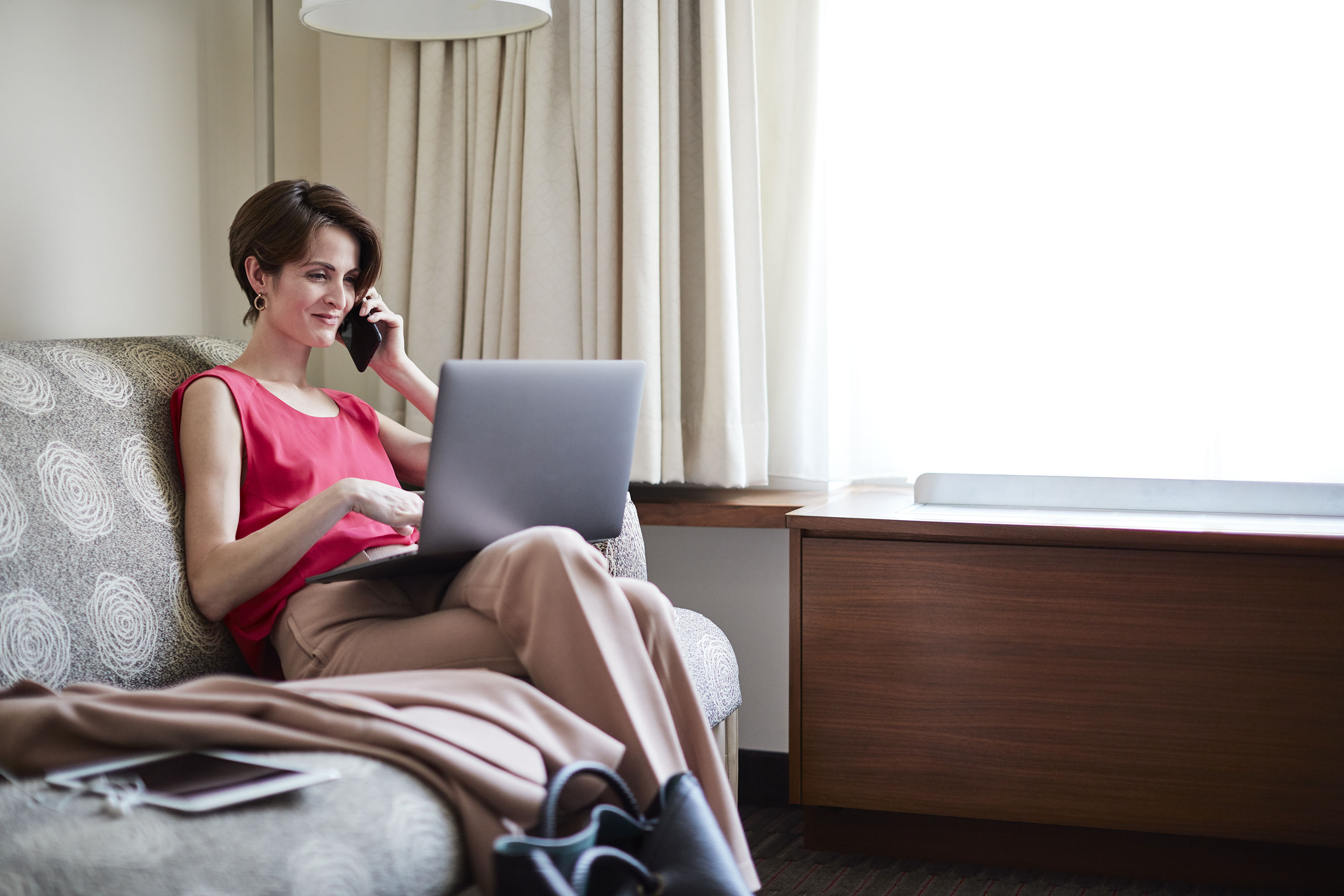 A woman sitting on a sofa in a room, using a laptop and talking on the phone.