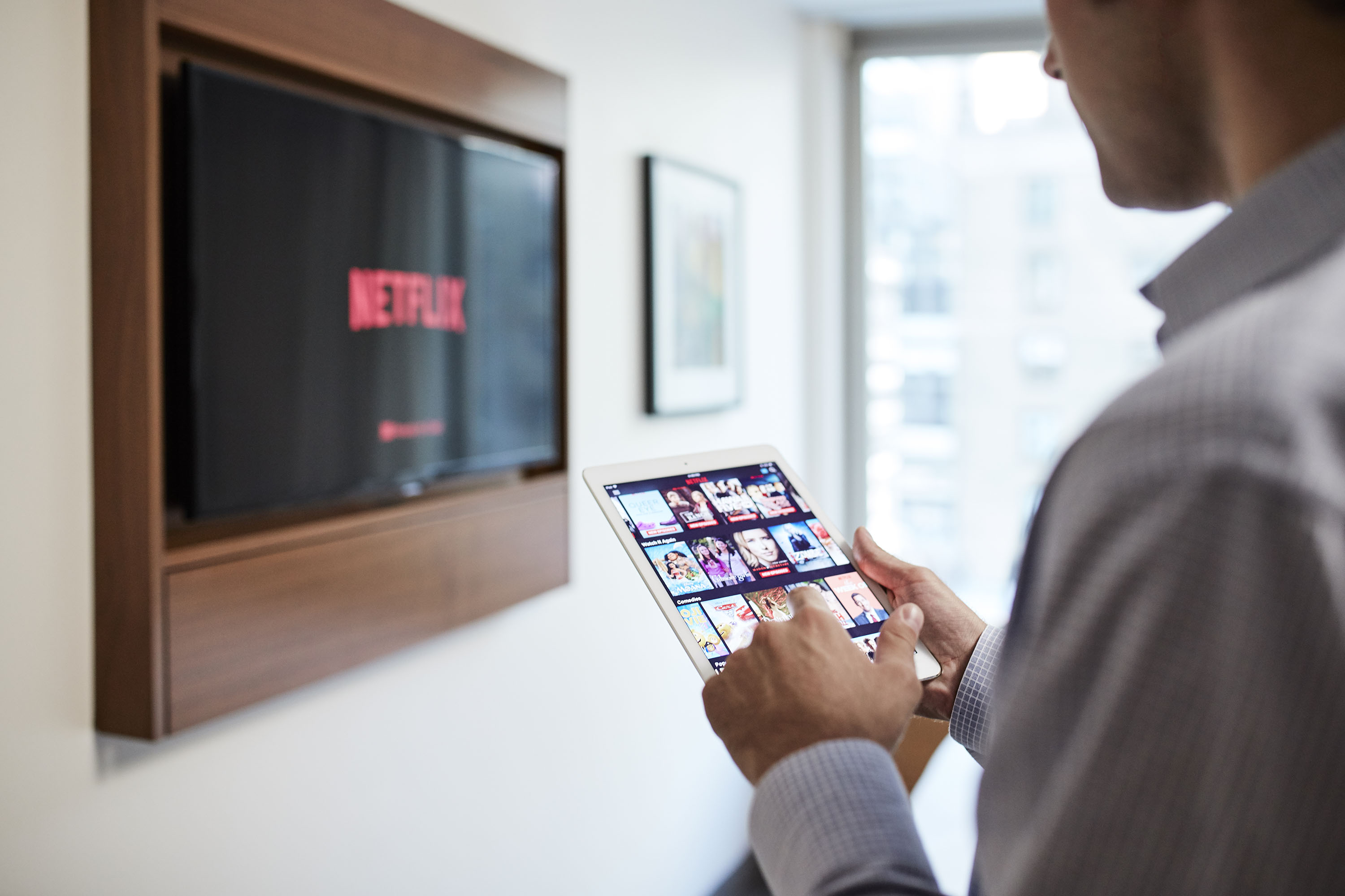 A man holding a tablet in front of a wall-mounted TV displaying Netflix.