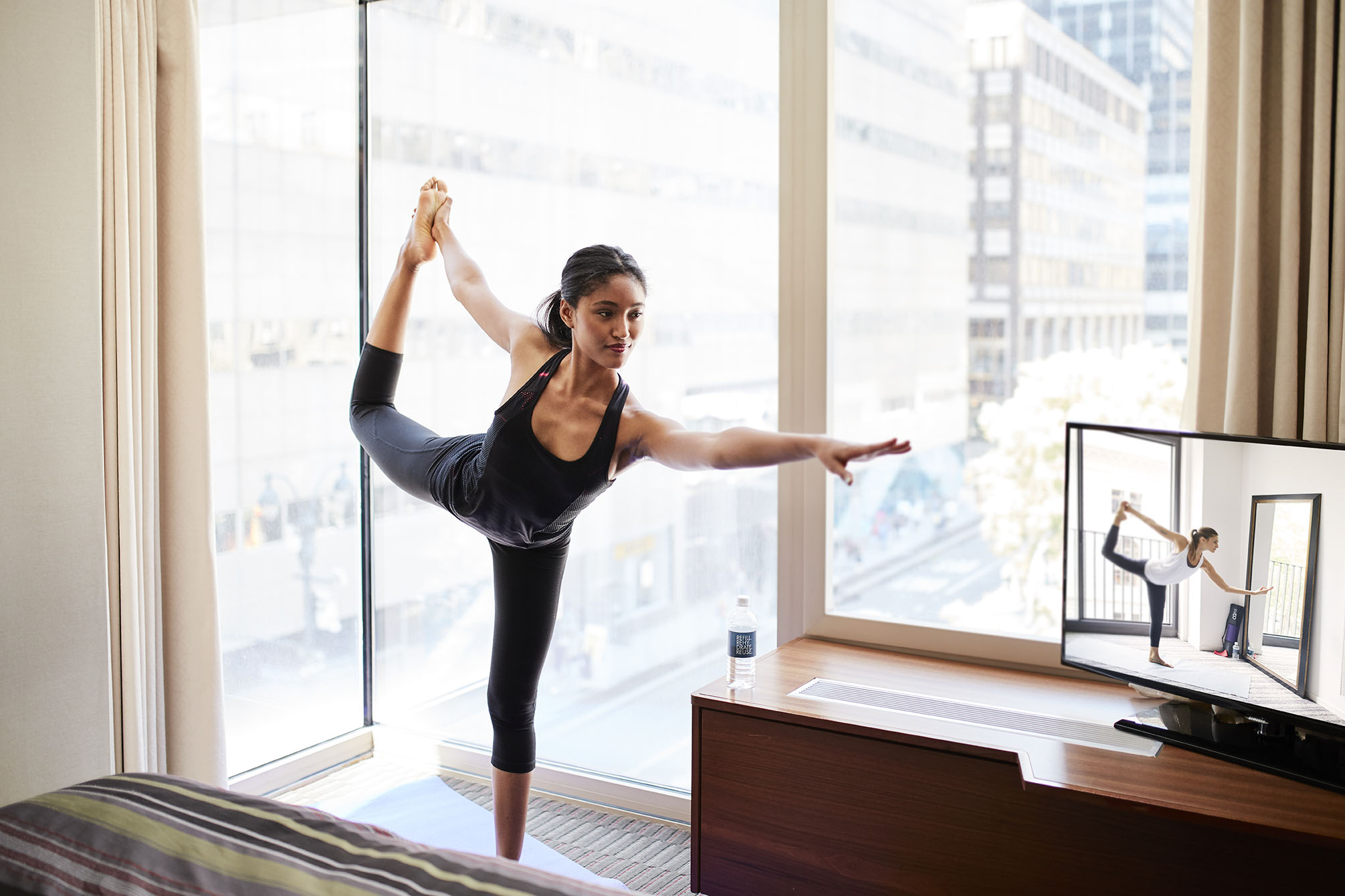 A woman in black workout clothes practicing yoga in a hotel room with a city view.