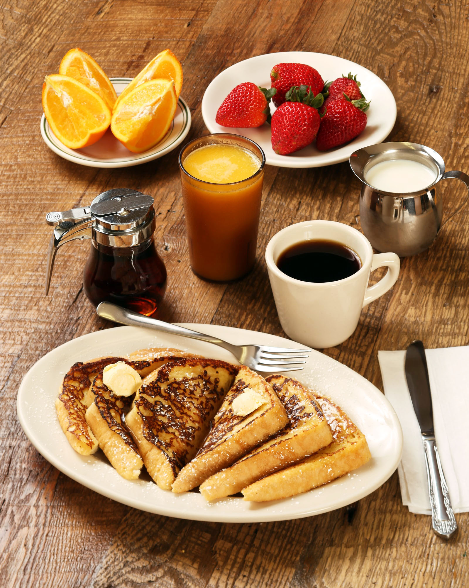 A breakfast spread featuring French toast, strawberries, oranges, coffee, and syrup on a wooden table.